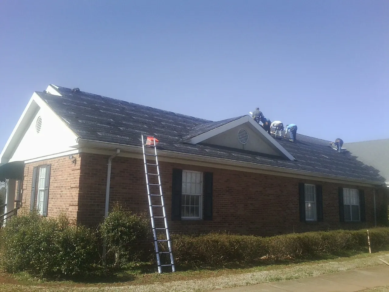 Crew tearing shingles off a brick ranch roof with a ladder set — Metal Roof Repair work by our crew in Harwood